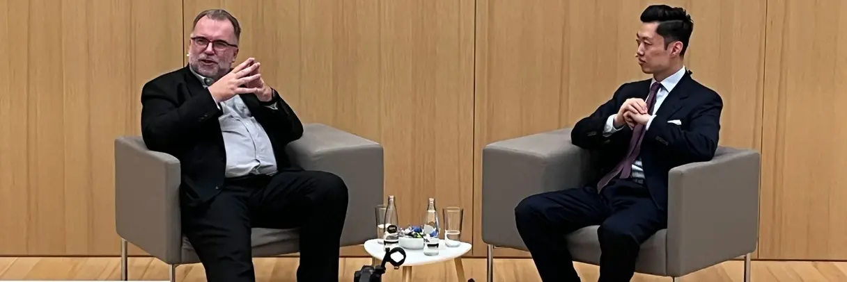 Two men in formal attire sitting in chairs, having a dialogue.