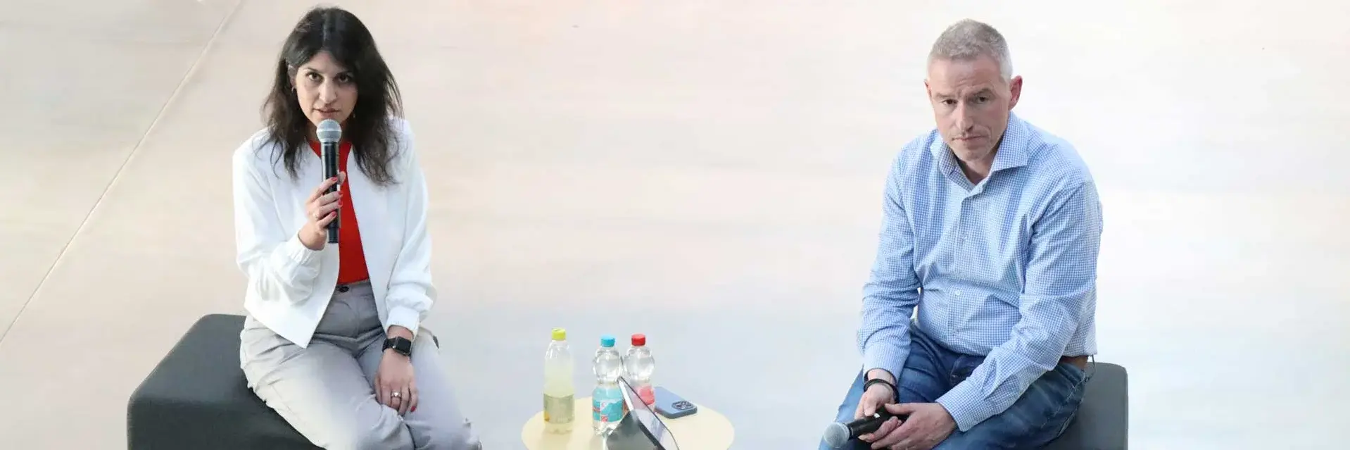 Two individuals seated on a stage, each with a microphone, engaged in conversation or discussion.