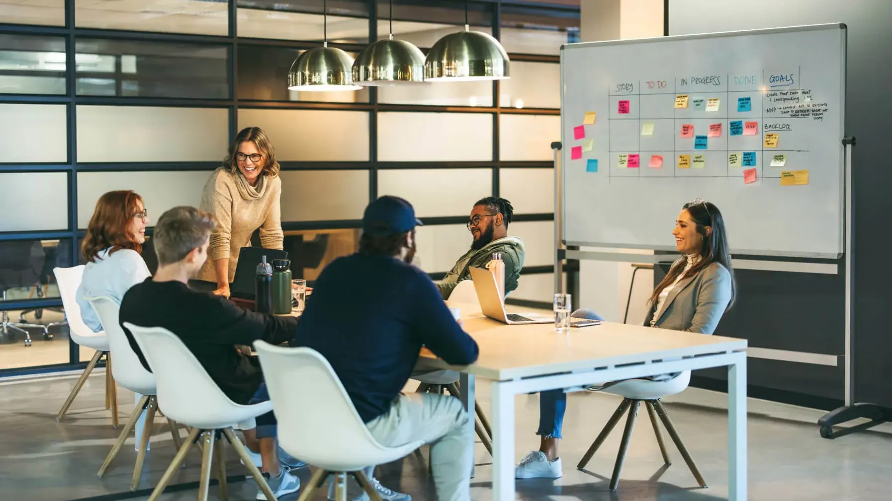 A diverse group of six people in a modern office setting participate in a meeting around a table. A woman stands near a whiteboard covered with colorful sticky notes. The atmosphere is collaborative and engaged.