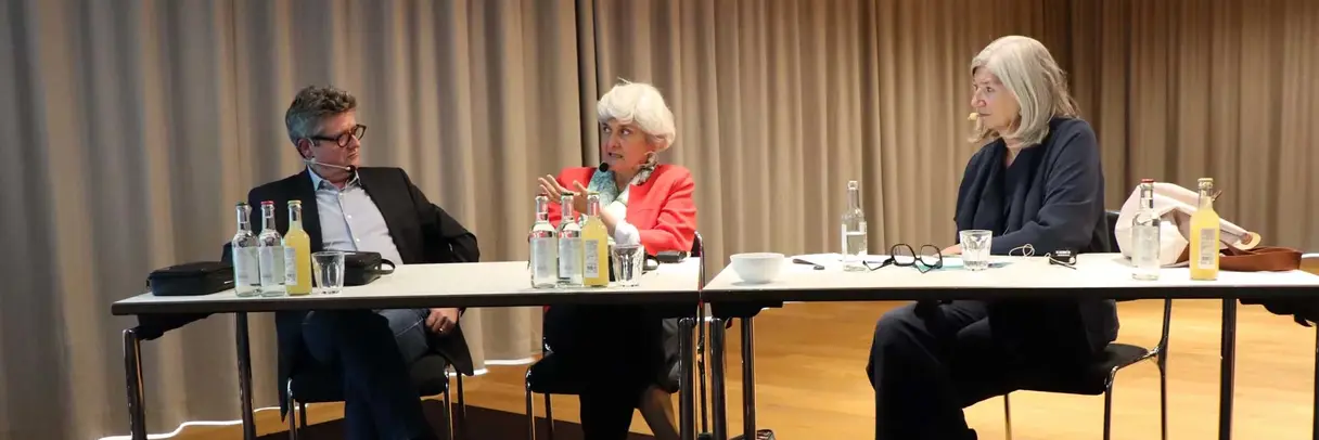 [Translate to German:] a woman und two men sitting behind a table, talking to each other